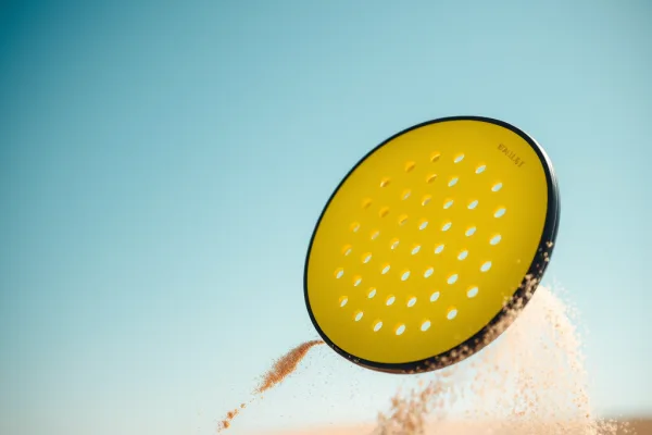 Bóng Pickleball màu vàng bay ngoài trời với 40 lỗ rõ ràng, có hiệu ứng gió và cát xung quanh, phong cách nhiếp ảnh khoa học.