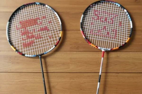 Hai cây vợt cầu lông, một cây chính hãng và một cây nhái, được đặt cạnh nhau trên mặt bàn gỗ.